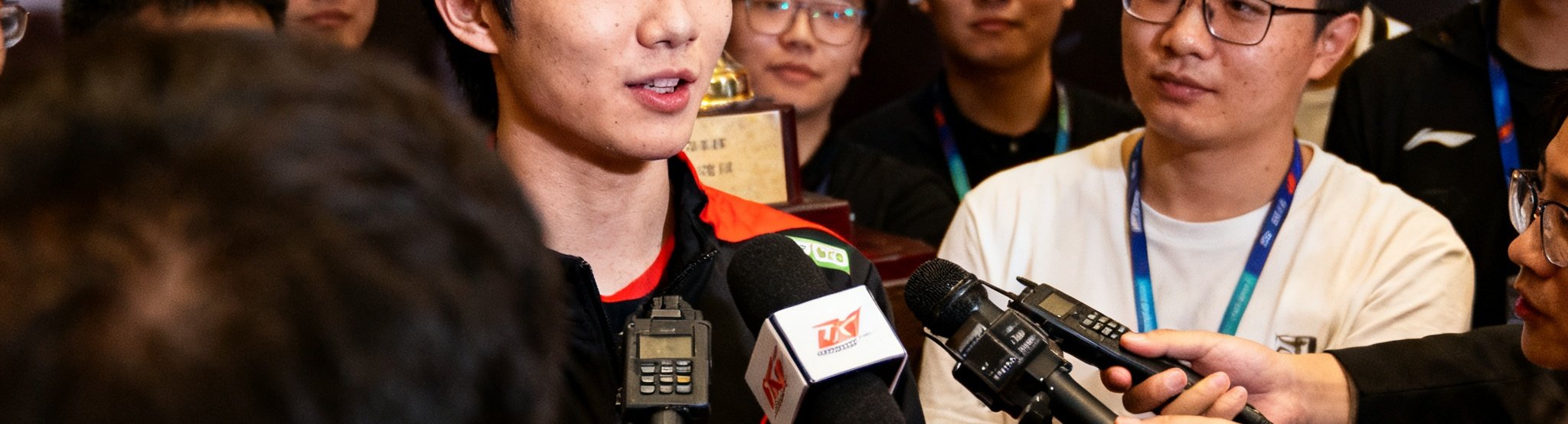 冠军选手在赛后采访区接受媒体专访，身后是熠熠生辉的冠军奖杯，记者们举着录音设备记录选手的真实感言